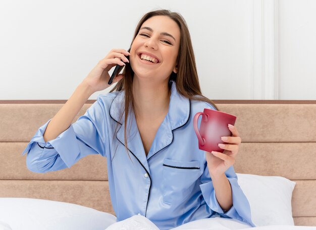 Belle jeune femme en pyjama bleu assis sur le lit avec une tasse de café parlant au téléphone mobile souriant joyeusement à l'intérieur de la chambre sur fond clair