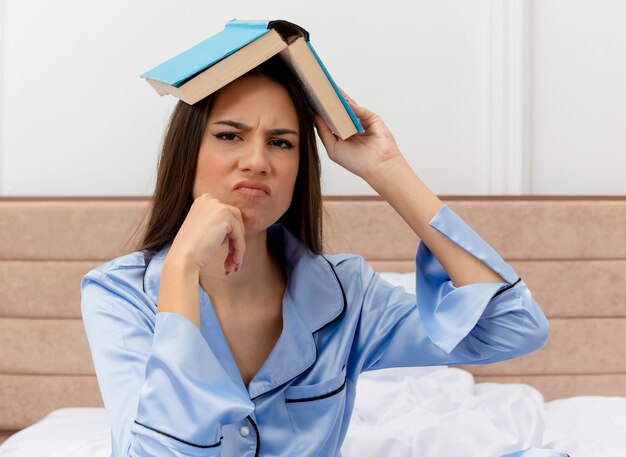 Belle jeune femme en pyjama bleu assis sur le lit tenant un livre au-dessus de sa tête regardant la caméra étant mécontent de l'intérieur de la chambre sur fond clair