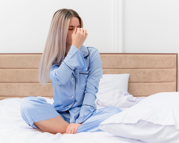 Belle jeune femme en pyjama bleu assis sur le lit en touchant son nez entre les yeux fermés fatigués et ennuyés dans l'intérieur de la chambre sur fond clair