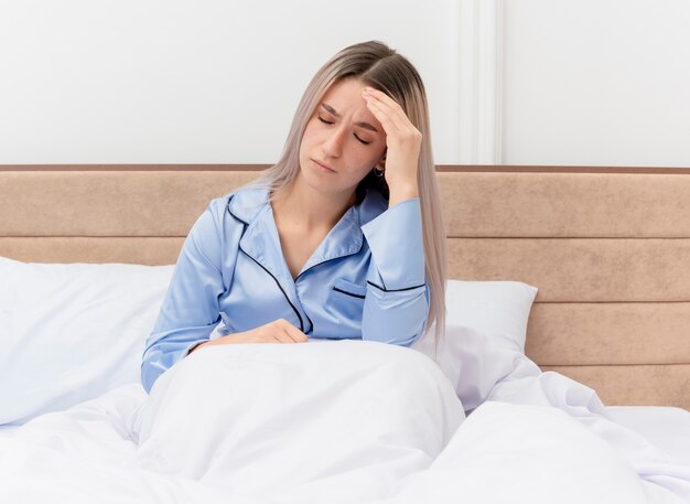 Belle jeune femme en pyjama bleu assise dans son lit touchant sa tête ayant des maux de tête à l'intérieur de la chambre
