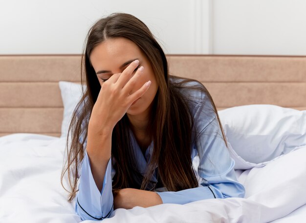 Belle jeune femme en pyjama bleu portant sur le lit avec oreiller bâillement sensation de fatigue matinale à l'intérieur de la chambre sur fond clair