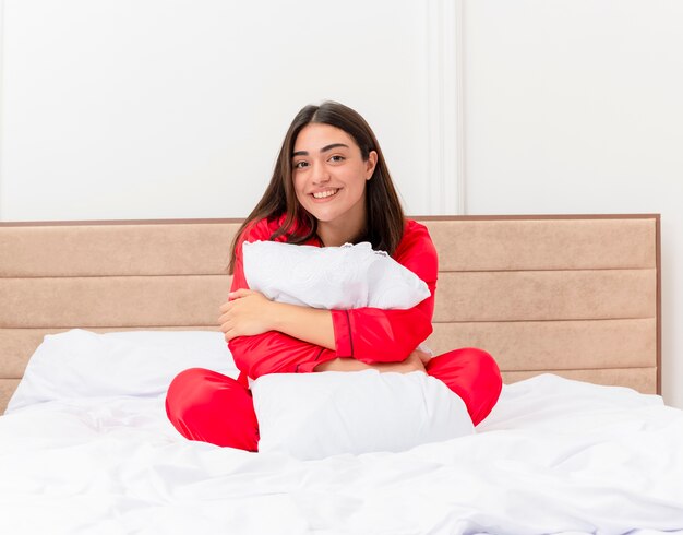 Belle jeune femme en pyjama rouge assis dans son lit étreignant l'oreiller lookign à la caméra souriant heureux et positif profitant du week-end à l'intérieur de la chambre