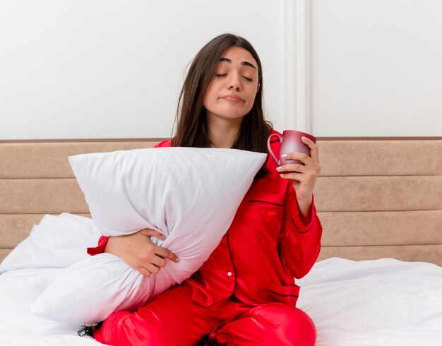 Belle jeune femme en pyjama rouge assis dans son lit avec oreiller tenant une tasse à la fatigue du matin sentiment fatigué dans l'intérieur de la chambre sur fond clair