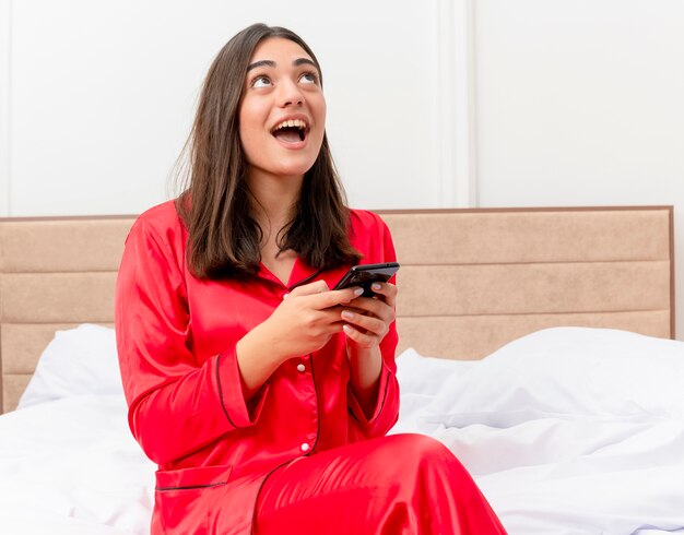 Belle jeune femme en pyjama rouge assis sur le lit à l'aide de smartphone à la recherche de sourire avec un visage heureux dans l'intérieur de la chambre sur fond clair