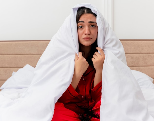 Photo gratuite belle jeune femme en pyjama rouge assis sur le lit enveloppant dans une couverture à la recherche de mal avec visage malheureux à l'intérieur de la chambre sur fond clair