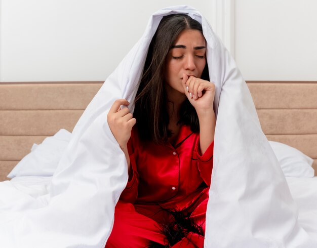 Belle jeune femme en pyjama rouge assis sur le lit enveloppant dans une couverture à la toux malade à l'intérieur de la chambre sur fond clair