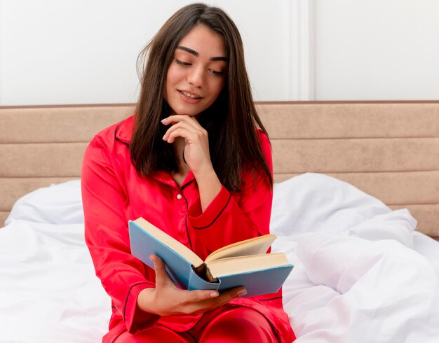 Belle jeune femme en pyjama rouge assis sur le lit avec lecture de livre heureux et positif à l'intérieur de la chambre sur fond clair