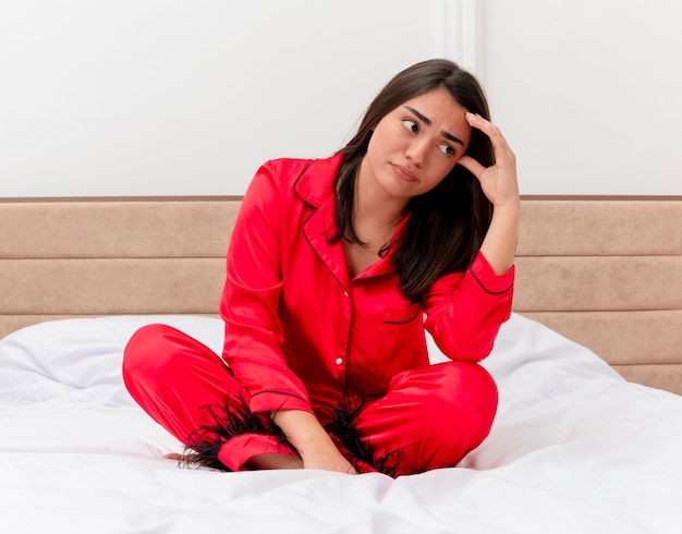 Belle jeune femme en pyjama rouge assis sur le lit lookign côté perplexe et mécontent à l'intérieur de la chambre sur fond clair