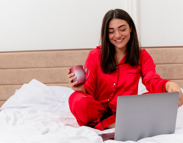 Belle jeune femme en pyjama rouge assis sur le lit avec ordinateur portable et tasse de café heureux et positif souriant joyeusement à l'intérieur de la chambre sur fond clair