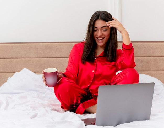 Belle jeune femme en pyjama rouge assis sur le lit avec ordinateur portable et tasse de café heureux et positif souriant joyeusement à l'intérieur de la chambre sur fond clair