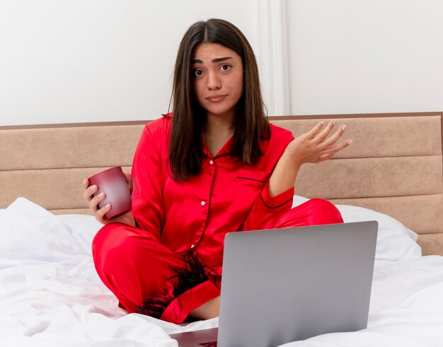 Belle jeune femme en pyjama rouge assis sur le lit avec un ordinateur portable et une tasse de café regardant la caméra confus étalant le bras sur le côté dans l'intérieur de la chambre sur fond clair