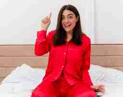 Photo gratuite belle jeune femme en pyjama rouge assis sur le lit en regardant la caméra heureux souriant montrant l'index ayant une nouvelle idée dans l'intérieur de la chambre sur fond clair