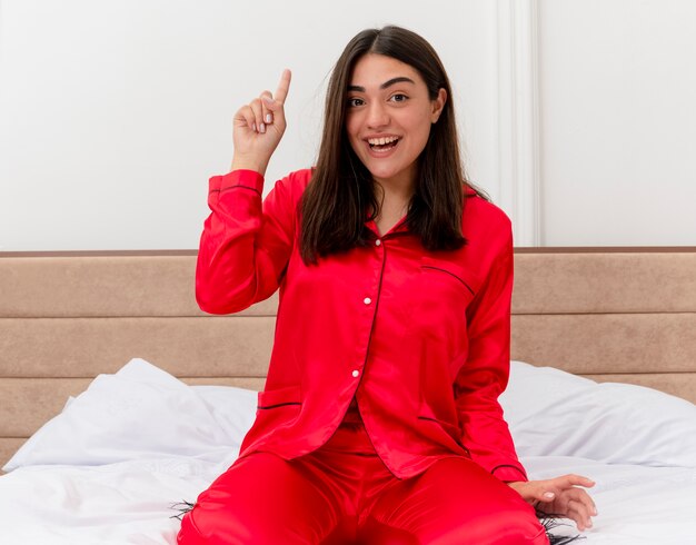 Belle jeune femme en pyjama rouge assis sur le lit en regardant la caméra heureux souriant montrant l'index ayant une nouvelle idée dans l'intérieur de la chambre sur fond clair