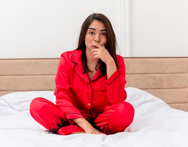 Belle jeune femme en pyjama rouge assis sur le lit en regardant la caméra avec un visage sérieux à l'intérieur de la chambre sur fond clair