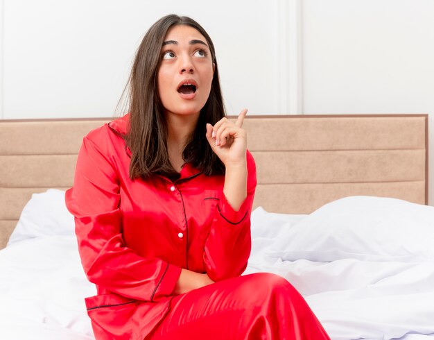 Belle jeune femme en pyjama rouge assis sur le lit regardant excité montrant l'index ayant une nouvelle idée dans l'intérieur de la chambre sur fond clair