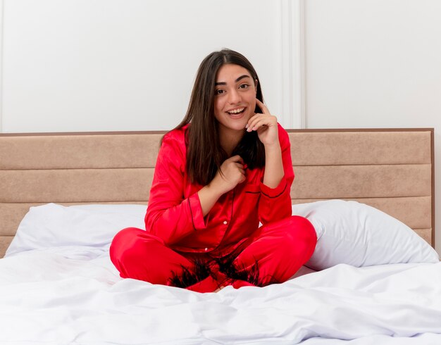 Belle jeune femme en pyjama rouge assis sur le lit relaxant souriant avec un visage heureux à l'intérieur de la chambre