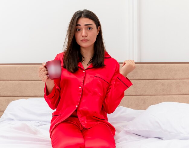 Belle jeune femme en pyjama rouge assis sur le lit avec une tasse de café regardant la caméra étant confus et très anxieux dans l'intérieur de la chambre sur fond clair