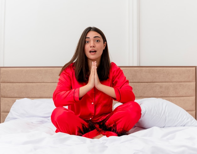 Belle jeune femme en pyjama rouge assis sur le lit tenant les paumes ensemble comme prier ou namaste geste émotionnel et inquiet à l'intérieur de la chambre sur fond clair