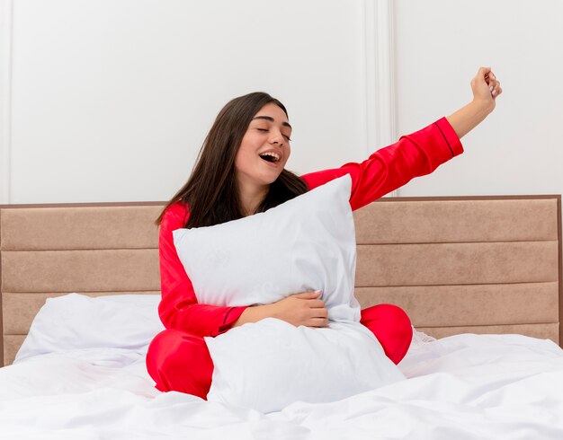 Belle jeune femme en pyjama rouge assise dans son lit avec un oreiller heureux et excité de profiter du week-end à l'intérieur de la chambre
