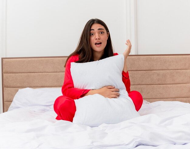 Belle jeune femme en pyjama rouge assise dans son lit avec un oreiller surprise et étonnée à l'intérieur de la chambre
