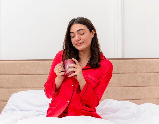 Belle jeune femme en pyjama rouge assise sur le lit avec une tasse de café les yeux fermés ressentant des émotions positives à l'intérieur de la chambre