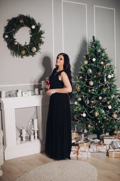 Belle jeune femme de race blanche aux longs cheveux noirs en longue robe noire se tient près de l'arbre de Noël avant le dîner
