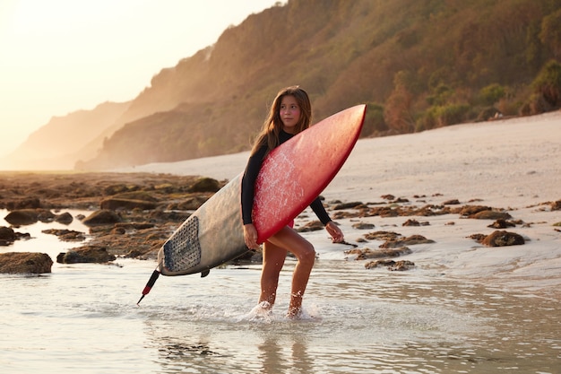 Belle jeune femme de race blanche bronzée porte planche de surf, se promène sur l'eau près de la plage de sable
