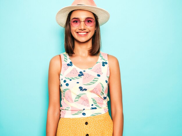 Belle jeune femme regardant la caméra au chapeau. Fille à la mode en T-shirt blanc d'été décontracté et jupe jaune à lunettes de soleil rondes. La femelle positive montre des émotions faciales. Modèle drôle isolé sur bleu