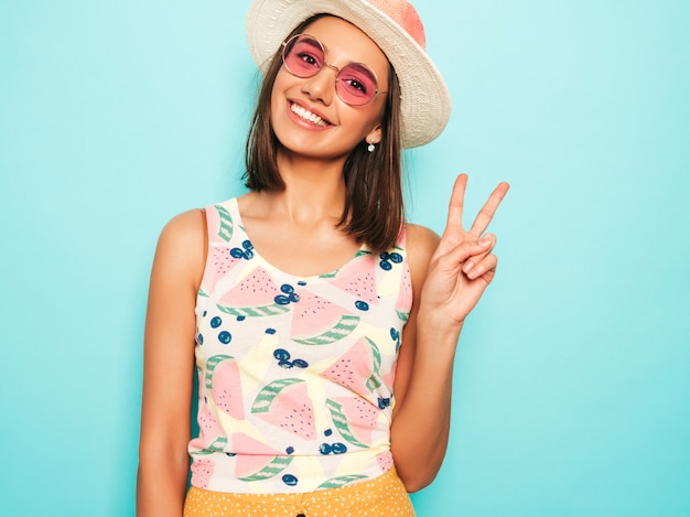 Photo gratuite belle jeune femme regardant la caméra au chapeau. fille à la mode en t-shirt blanc d'été décontracté et jupe jaune à lunettes de soleil rondes. la femelle positive montre des émotions faciales. montre le signe de la paix