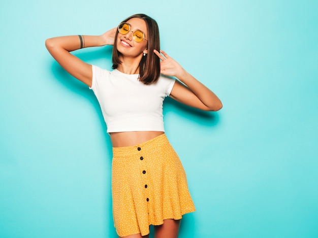 Belle jeune femme regardant la caméra. Fille à la mode en T-shirt blanc d'été décontracté et jupe jaune à lunettes de soleil rondes. La femelle positive montre des émotions faciales. Modèle drôle isolé sur bleu