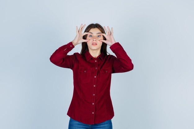 Belle jeune femme regardant à travers les doigts en chemise et l'air concentré.