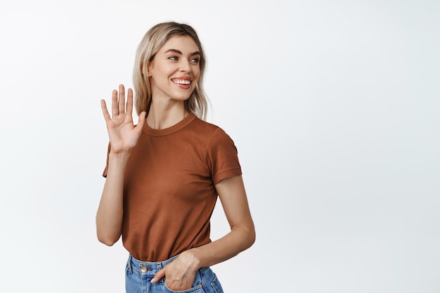 Belle jeune femme regarde derrière son épaule et agite la main pour dire bonjour en souriant et en regardant amicalement debout sur fond blanc