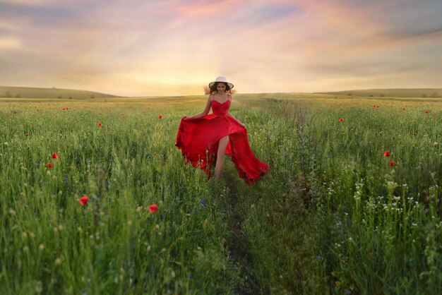 Belle jeune femme en robe rouge et chapeau blanc se promène sur le terrain avec des coquelicots