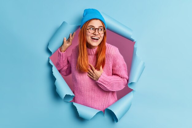 Belle jeune femme rousse garde la main sur la poitrine et regarde positivement de côté remarque que la chose drôle porte chapeau rose pull tricoté traverse le trou de papier