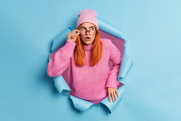 Belle jeune femme rousse surprise garde la bouche ouverte voit quelque chose d'incroyable étonne par une offre inattendue porte un chapeau rose et un pull traverse du papier bleu