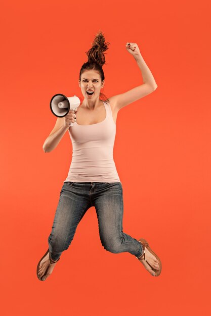 Belle jeune femme sautant avec mégaphone isolé sur fond rouge. Fille Runnin en mouvement ou en mouvement. Concept d'émotions humaines et d'expressions faciales