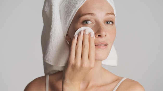 Belle jeune femme avec une serviette sur la tête nettoyé le visage avec une éponge de coton sur fond blanc. Notion de beauté