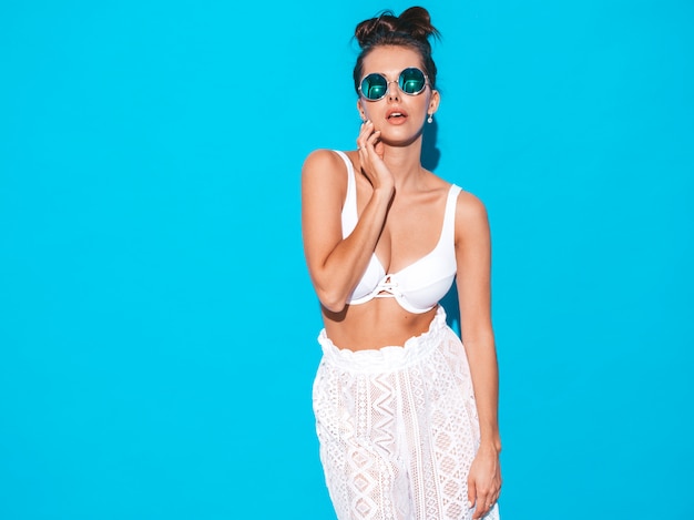 Belle jeune femme sexy avec une coiffure de goule. Fille branchée en pantalon blanc d'été décontracté et soutien-gorge en lunettes de soleil. Modèle chaud isolé sur bleu