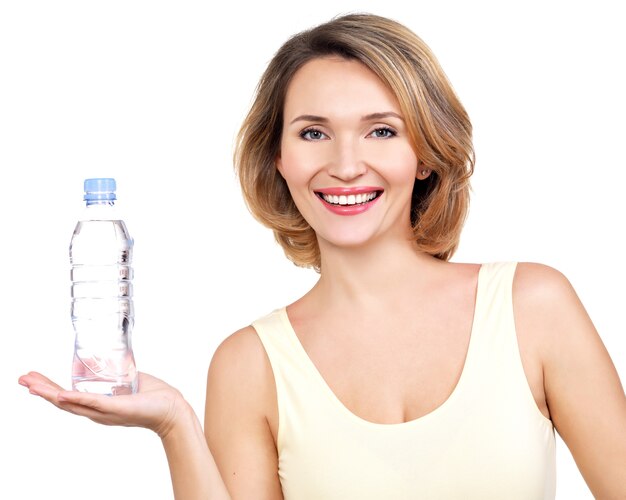 Belle jeune femme souriante avec une bouteille d'eau sur un mur blanc.