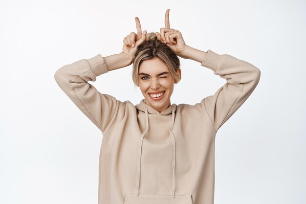 Belle jeune femme souriante avec un clin d'œil et montrant le geste des cornes de taureau debout dans un sweat à capuche beige sur fond blanc