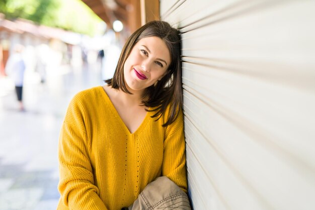 Belle jeune femme souriante confiante et joyeuse s'appuyant sur un mur en bois marchant dans la rue de la ville par une journée ensoleillée