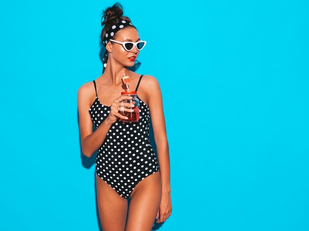 Belle jeune femme souriante sexy hipster en lunettes de soleil.Fille en maillot de bain pois d'été maillot de bain.Poser près du mur bleu, boire un cocktail frais