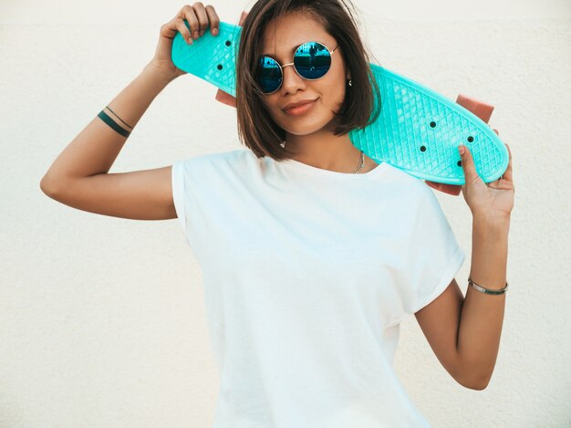 Belle jeune femme souriante sexy hipster en lunettes de soleil.Tendance fille en été T-shirt et short.Positive femelle avec bleu penny skateboard posant dans la rue près du mur blanc