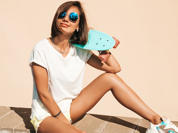 Belle jeune femme souriante sexy hipster en lunettes de soleil.Tendance fille en été T-shirt et shorts.Femme positive avec skateboard penny bleu posant sur le fond de la rue