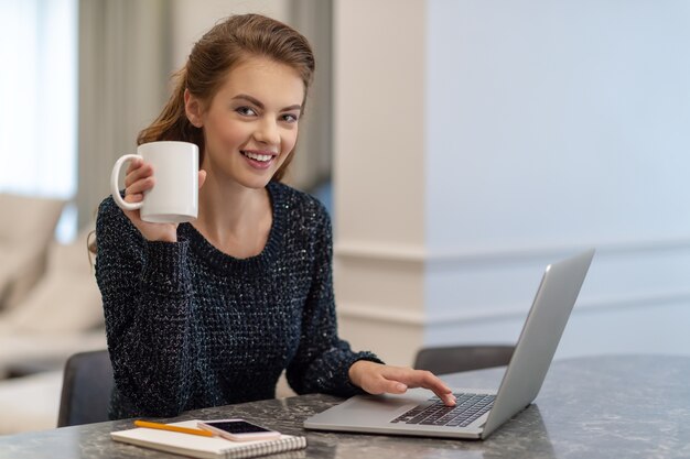 Belle jeune femme souriante travaillant sur ordinateur portable et boire du café alors qu'il était assis à la maison