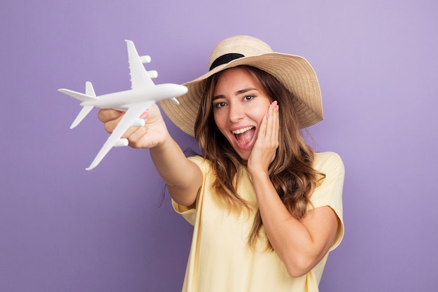 Belle jeune femme en t-shirt beige et chapeau d'été tenant un jouet regardant la caméra avion heureux et excité debout sur fond violet