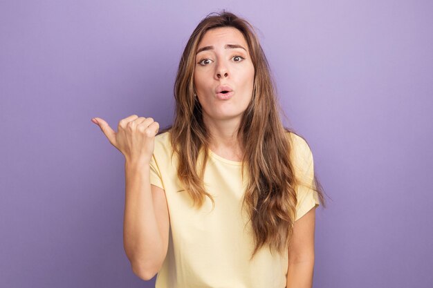 Belle jeune femme en t-shirt beige regardant la caméra confuse et surprise pointant avec le pouce en arrière