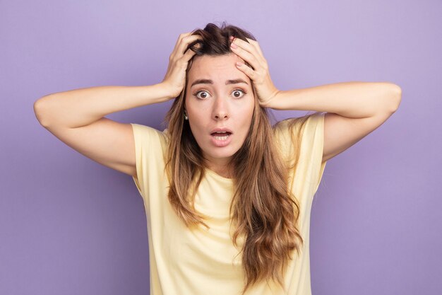 Belle jeune femme en t-shirt beige regardant la caméra étonnée avec les mains sur la tête