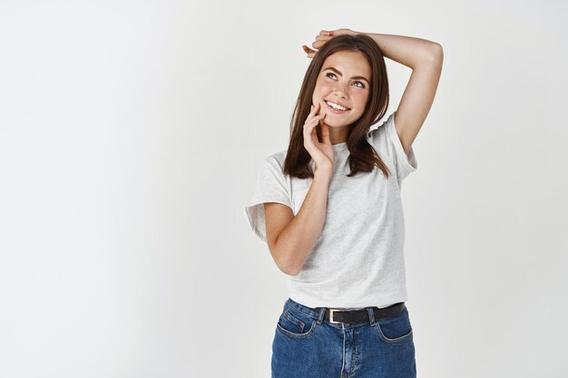 Belle jeune femme en t-shirt blanc rêveuse en levant, posant sur le mur du studio.