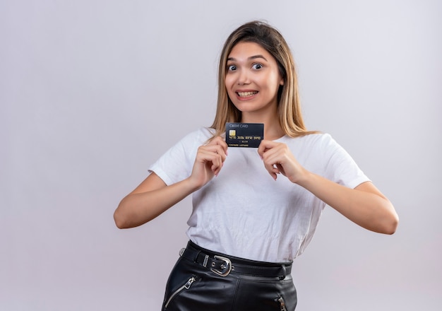 Une belle jeune femme en t-shirt blanc souriant tout en montrant la carte de crédit sur un mur blanc